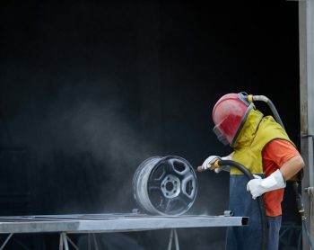 Caucasian man dressed in special protective equipment using sandblaster for cleaning metal details at work. Concept of people and repairing.